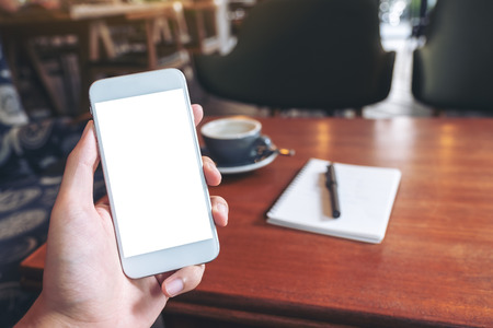 Mockup image of hand holding white mobile phone with blank desktop screen with coffee cup and notebook on wooden table in cafeの写真素材