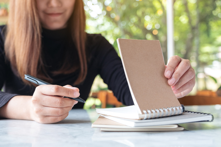 Closeup image of a business woman holding and writing on blank notebook on table with green nature backgroundの写真素材