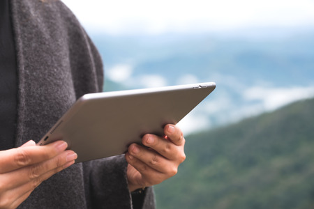 Closeup image of a woman holding and using tablet pc with green mountains on foggy day with blue sky...