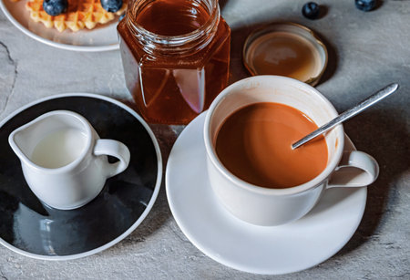 Coffee with milk in a cup and a jar of honey nearby.の写真素材