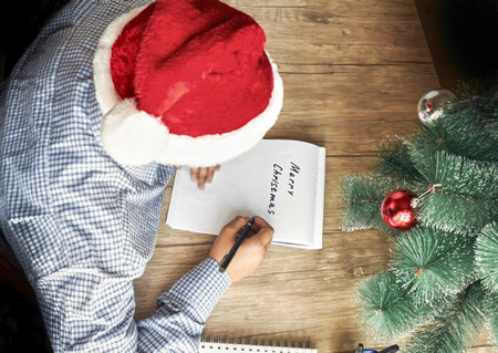 A guy in a Santa hat writes Merry Christmas on a white piece of paperの写真素材