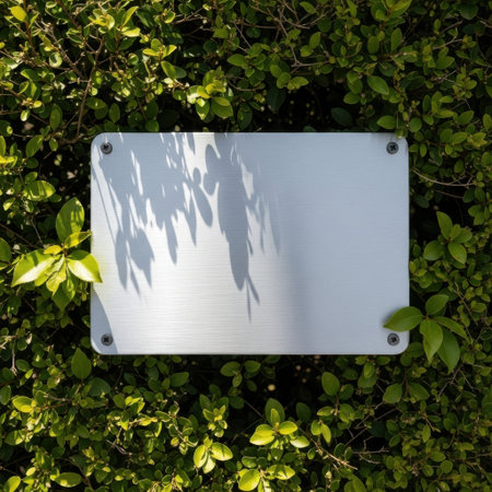 A blank, rectangular metal plaque is mounted securely to a vibrant green hedge. Sunlight glints off the reflective surface, casting abstract patterns of light and shadow from nearby foliage.の素材
