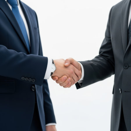 A close-up view of two professionals in dark suits and ties shaking hands, presented isolated on a clean white background.の素材