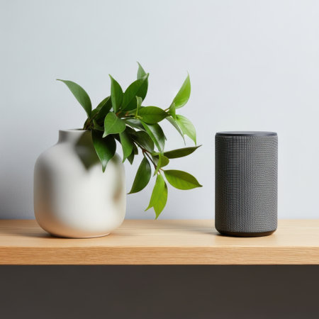A stylish white vase holds vibrant green leaves next to a dark gray cylindrical smart device on a wooden surface.の素材
