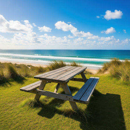 A rustic wooden picnic table sits on a grassy dune overlooking a tranquil beach with gentle waves under a vibrant blue sky filled with fluffy white clouds.の素材