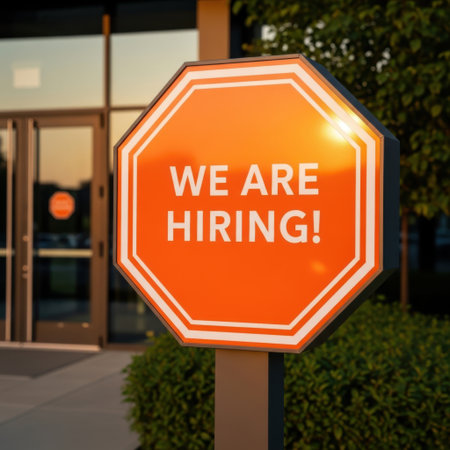 A vibrant orange octagonal sign with the message 'We Are Hiring!' is mounted on a pole. The sign is illuminated, attracting attention to the employment opportunity outside a building.の素材