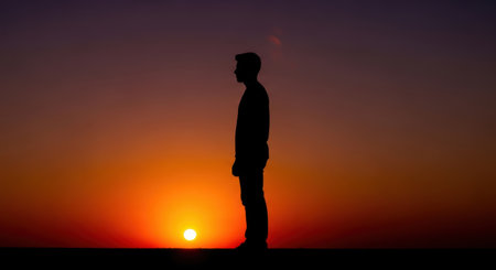 A solitary person is depicted as a silhouette against a breathtaking sunset. The sky glows with intense orange and red tones, creating a dramatic and atmospheric backdrop for the contemplative figure.の素材