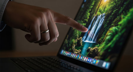 A person is using a laptop to select a scenic image of a waterfall. The laptop screen displays a vibrant waterfall surrounded by lush greenery. The user's hand is pointing at the waterfall image, indicating a selection or interaction.の素材