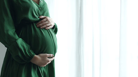 Pregnant woman in a green dress gently holding her belly by a window