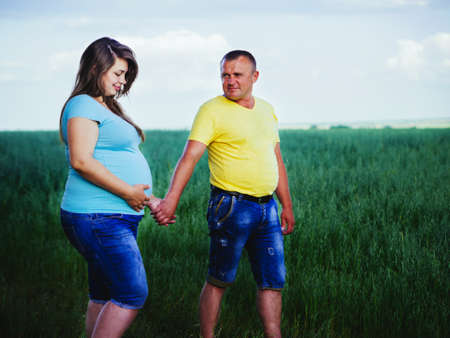 Smiling beautiful pregnant woman looking at her belly and holds hand of her husband outdoors. Casual couple holding hands and walking in a meadow.の写真素材