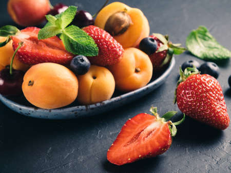 Closeup view of fruits and berries on dark background. Heap of fresh strawberries, blueberries, apricot and mint leaves in trendy plate. Focus on strawberry. Healthy food,superfood,diet,detox concept.の写真素材