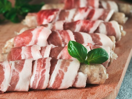 Cold prepared to grill pigs sausages wrapped in bacon on plate. Raw sausages wrapped in bacon served fresh green basil leaves on wooden cutting board. Shallow DOF. Copy space for text.の写真素材