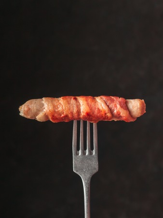 Ready-to-eat pigs sausages wrapped in bacon on rustic fork. Fried savory sausages wrapped in bacon isolated on black background. Copy space for text.の写真素材