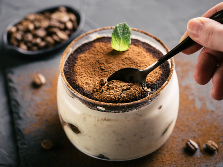 High angle view of tiramisu in glass cup on stone plate over black concrete background, Hand with teaspoon eats tiramisu. Copy space.の写真素材
