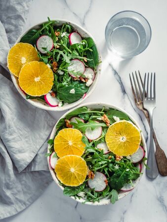 Vegan salad bowl with oranges, spinach, arugula, radish, nut. Top view. Vegan breakfast, vegetarian food, diet concept. Two bowls with salad on white marble tabletop. Verticalの写真素材
