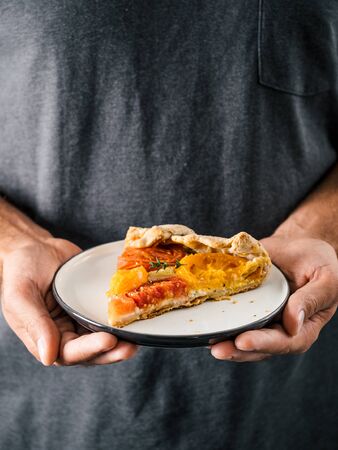 Hands takes plate with portion piece of pie. Savory fresh homemade tomato tart or galette. Healthy appetiezer - whole wheat or rye-wheat pie with tomatoes,cheese. Vertical. Copy space for textの写真素材