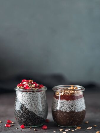 Two chia pudding in glass jars on dark table. Two colors charcoal chia puding with pomegranate, almond, hemp seeds. Chocolate chia pudding with jam and granola. Copy space. Healthy overnight breakfastの写真素材