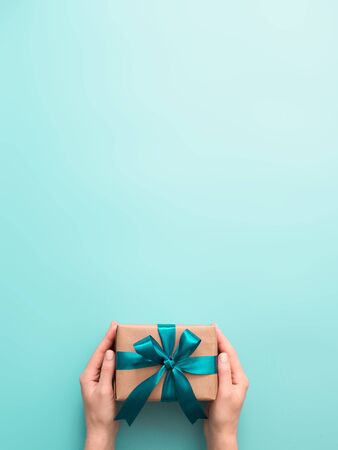 Female hands hold gift box on turquoise blue background, copy space. Caucasian girl hands holding gift box in craft wrapping paper with green satin ribbon. Christmas, New Year or Birthday presentの写真素材