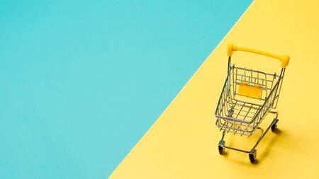 Empty miniature shopping cart on blue and yellow background. Toy trolley on bright colorful background, copy space for text or design.の写真素材