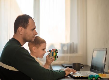 Four year old boy play and father at the table with notebook. Home working at coronavirus quarantine isolation period. Working among children, working remote concept. Sunset lightの写真素材