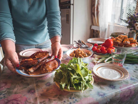 Woman sets kitchen table in country house. Woman hold plate with smoked fish on the table. Table setting in country house with wild flowers bouqet. Cottagecore and farmcore concept. Authentic shotの写真素材