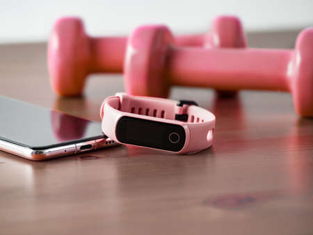 Fitness tracker on smartphone and pink colored dumb-bell on wooden tabletop. Pink female pedometer or wearable device on smartphone with infinity display, copy spaceの写真素材
