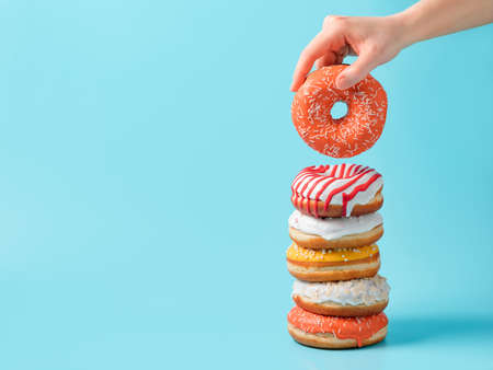 Stack of assorted donuts and one donut in female hand on blue background. Many colorful glazed doughnut with sprinkles stacked with copy spaceの写真素材