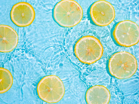 Lemon slices in clean transparent water over blue background with copy space. Water splashing on blue water surface in sunlight. Top view or flatlay. Summer, vacation, healthy eating conceptの写真素材