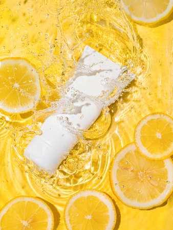 Cosmetic empty white tube in clean transparent water with lemon slices over yellow background. Water splashing on surface, sunlight. Vertical flatlay,copy space. Beauty mockup for cream or moisturizerの写真素材