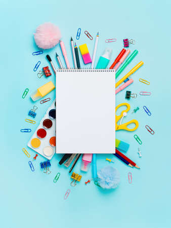 Notebook with stationary objects on blue background. Empty white paper sheet of notepad or sketchbook over shool supplies, isolated on blue. Vertical. Copy space for text or design. Top view, flat layの写真素材
