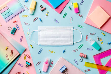 Protective medical mask on creative student desk with colorful modern school supplies on blue and pink duotone background. Top view. Flat lay. Copy space. Back to school covid19 conceptの写真素材