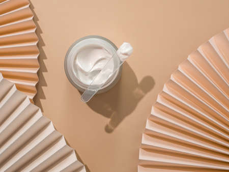 Pastel beige composition with cosmetic cream or moustirizer and fans. Open round glass jar with aesthetic swirls face cream and spatula. Top view or flat lay. Copy space. Cosmetic mockup.の写真素材