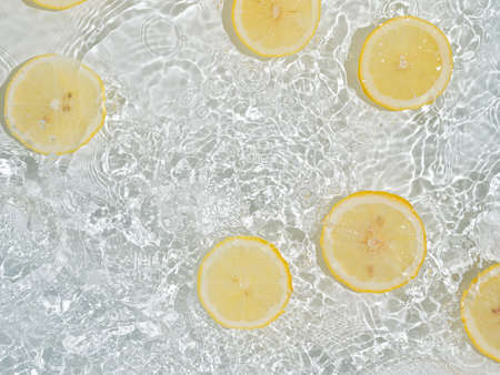 Lemon slices in clean transparent water over white background with copy space. Water splashing on water surface in sunlight. Top view or flatlay. Summer, vacation, healthy eating conceptの写真素材