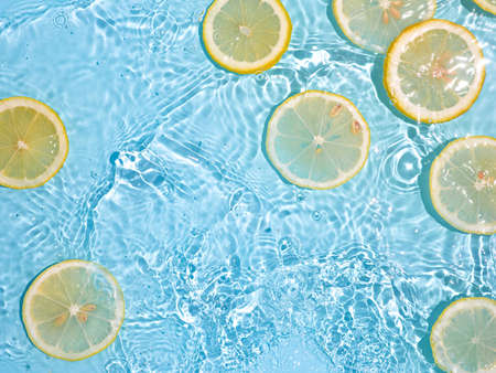 Lemon slices in clean transparent water over blue background with copy space. Water splashing on blue water surface in sunlight. Top view or flatlay. Summer, vacation, healthy eating conceptの写真素材