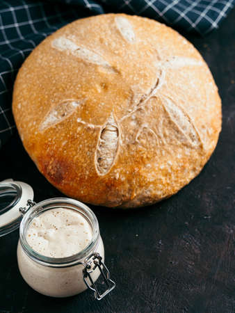Sourdoug starter and wheat sourdough bread. Wheat sour dough starter in glass jar and homemade round sourdough bread on black background, copy space. Home made sourdough bread making concept Vertical.の写真素材