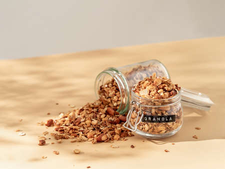 Homemade granola sprinkled of glass jar over pastel beige background. Aesthetic oat granola, beautiful shadows, champagne yellow background. Healthy breakfast concept. Copy spaceの写真素材