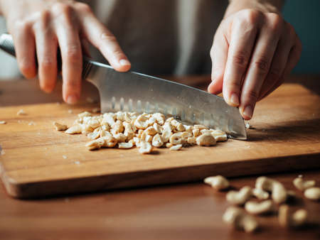 Female hands chopping cashew. Close up shot of process crushing raw unroasted cashew with large knife on wooden cutting board. Shallow DOF. Selective focusの写真素材