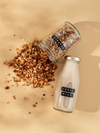 Granola and vegan milk. Healthy breakfast aesthetic concept. Homemade granola sprinkled of glass jar and vegan milk in glass bottle over champagne yellow or pastel beige background. Top view flat layの写真素材