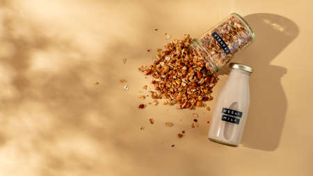 Granola and vegan milk. Healthy breakfast aesthetic concept. Homemade granola sprinkled of glass jar and vegan milk in glass bottle over champagne yellow or pastel beige background. Top view flat layの写真素材