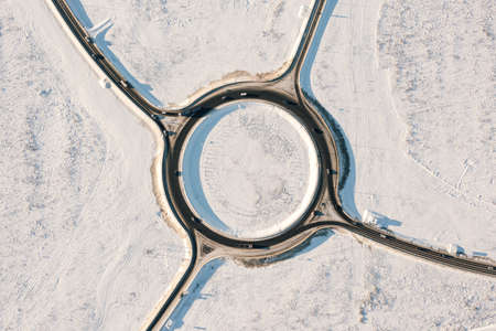 Aerial top view of circular traffic on winter road. Circle asphalt road with moving cars in sunny cold winter day. Car traffic on highway junction in winter seasonの写真素材