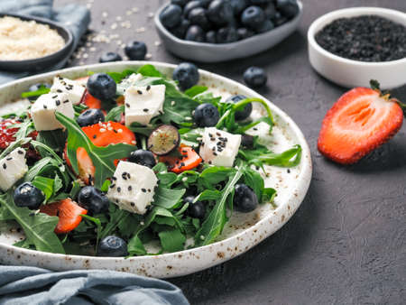 Salad with arugula, feta cheese and berries - strawberry, blueberry, in craft plate on black bacground. Summer salad idea and recipe for healthy vegetarian lunch, dinner. Copy space.の写真素材