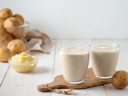 Potato milk in glasses on white wooden background. Pouring vegan milk in glass, with potato puree and potato tubers on background. Copy space. Home made potato milk made from boiled potatoesの写真素材