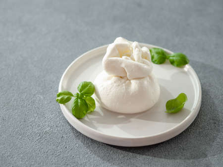 Fresh Italian cheese on plate. White ball of burrata or burratina cheese, mozzarella with cream filling. Soft white burrata cheese ball on round white plate, fresh basil. Gray background. Copy spaceの素材 [FY310188148902]