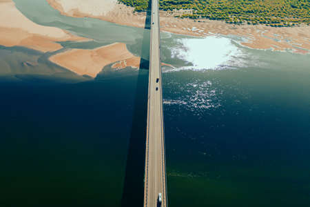 Aerial drone shot of cars driving on bridge over river and sandy riverside at sunset. Amazing aesthetic 4k top view shot with beautiful coast with dune sands. Drone aerial bridge with car trafficの写真素材