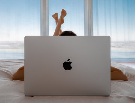 2022, Europe: Unrecognizable girl lying in bed and working behind macbook pro laptop on panoramic windows background. Young woman lying on bed and using mackbook on sea background with blue skyのeditorial素材
