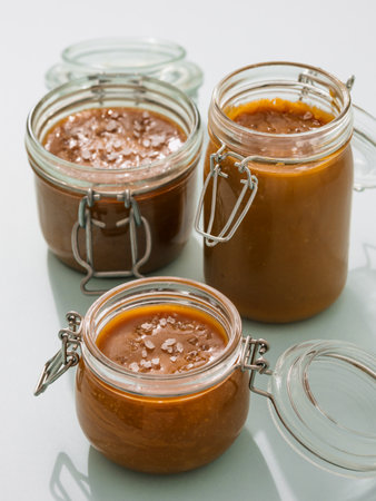 Salted caramel in glass jars. Brown caramel or condensed milk with sea salt crystalls, shoot in bright or hard light. Light neutral background. Verticalの写真素材