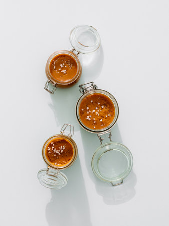 Salted caramel in glass jars, top view. Brown caramel or condensed milk with sea salt crystalls, shoot in bright or hard light. Light neutral background. Verticalの写真素材