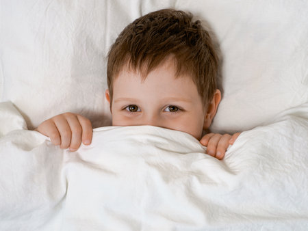 Mischief and play of young boy in bed before going to sleep. Four-year-old boy lies in bed on a pillow and looks out from under the coversの写真素材