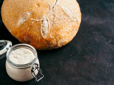Sourdoug starter and wheat sourdough bread. Wheat sour dough starter in glass jar and delicious homemade round sourdough bread on black background, copy space. Home made sourdough bread making conceptの写真素材