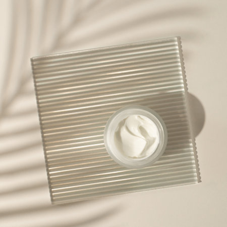 Cosmetic cream or moustirizer on transparent ribbed acrylic plate with tropical palm leaf shadows on pastel background. Open round glass jar with aesthetic swirls cream. Top view flatlay. Copy spaceの写真素材